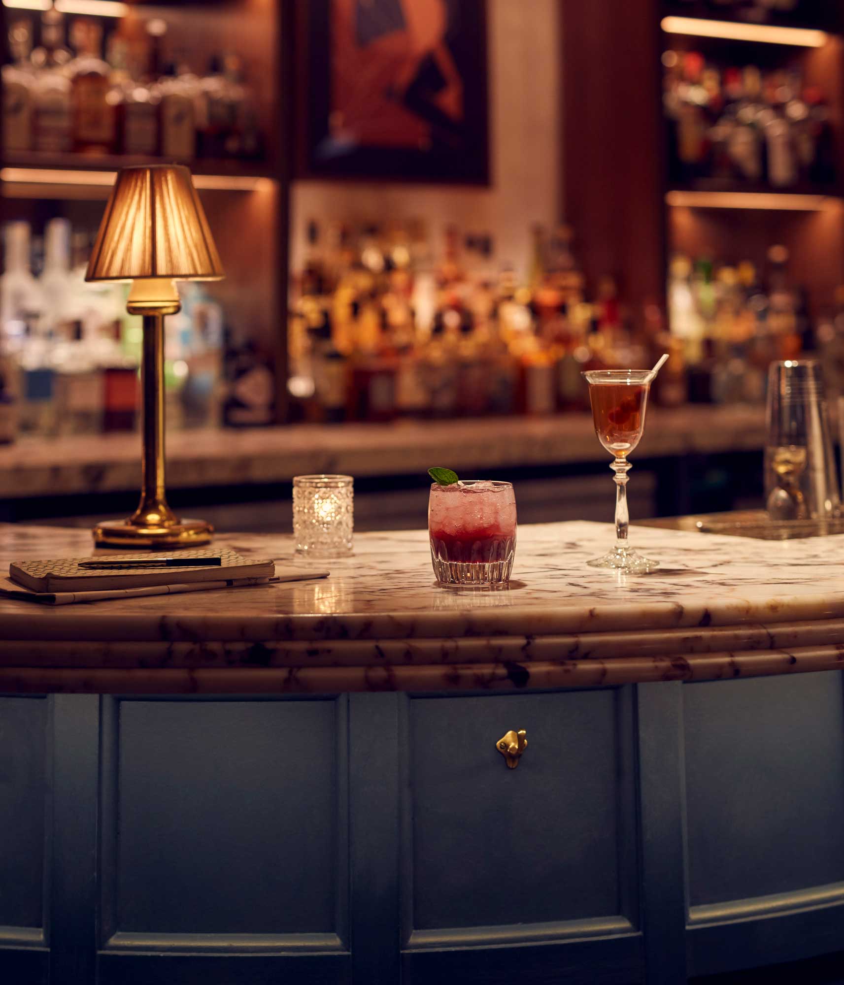 On the marbled bar top sits a dimly lit lamp, candle and two very different looking cocktails