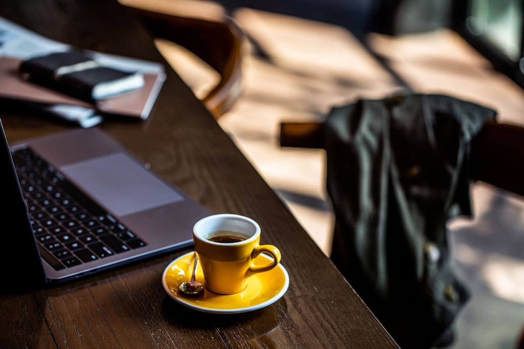 Long black in a bright yellow cup and saucer sitting beside a laptop