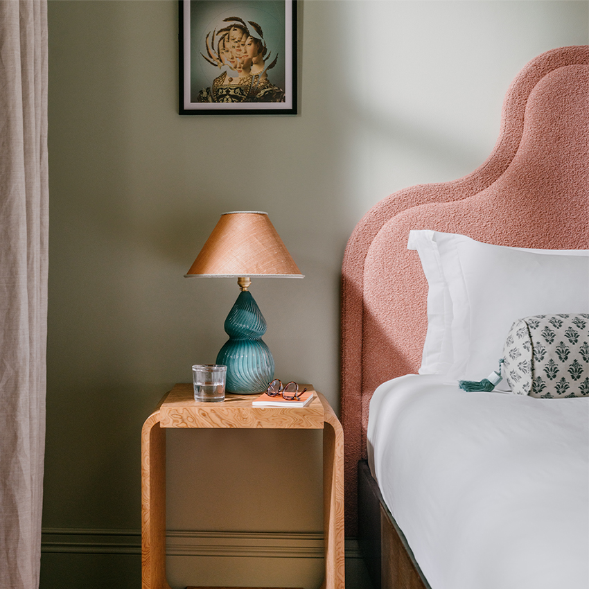 Natural sunlight illuminates a pink upholstered bed frame with a side table and a lamp beside it.