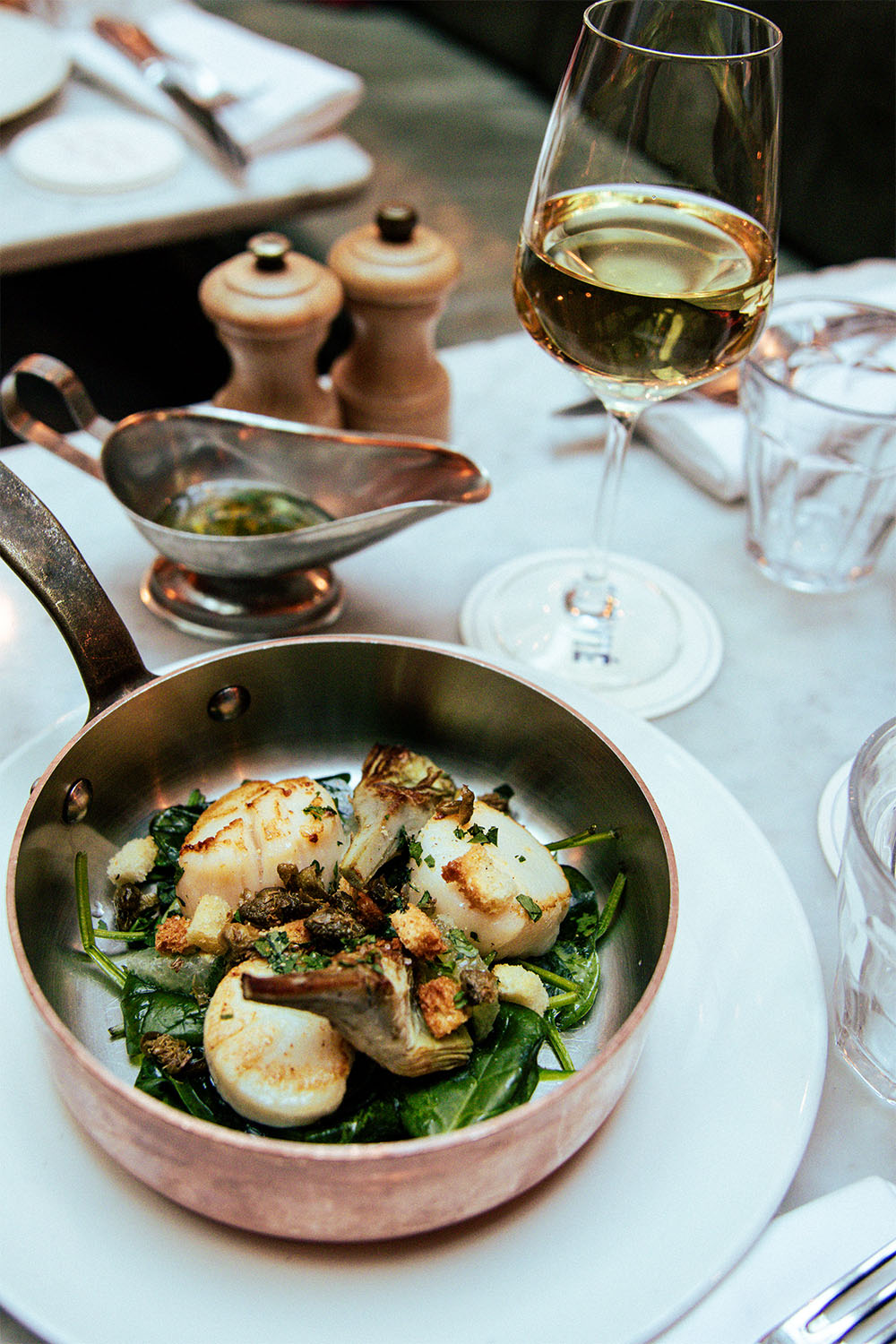 A table with a pan with scallop, mushrooms, spinach and herbs with a glass of white wine.