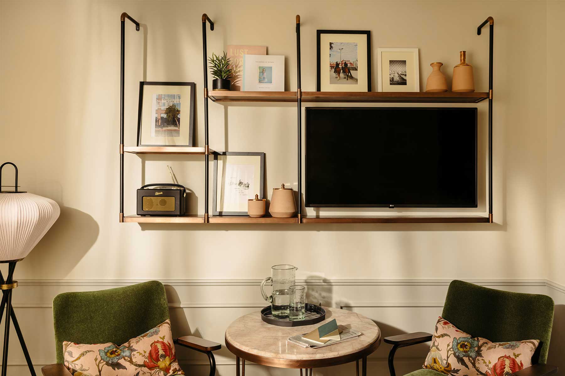 A shelving unit on the wall has artwork, vases, a radio and TV - it sits above two green lounge chairs and a table