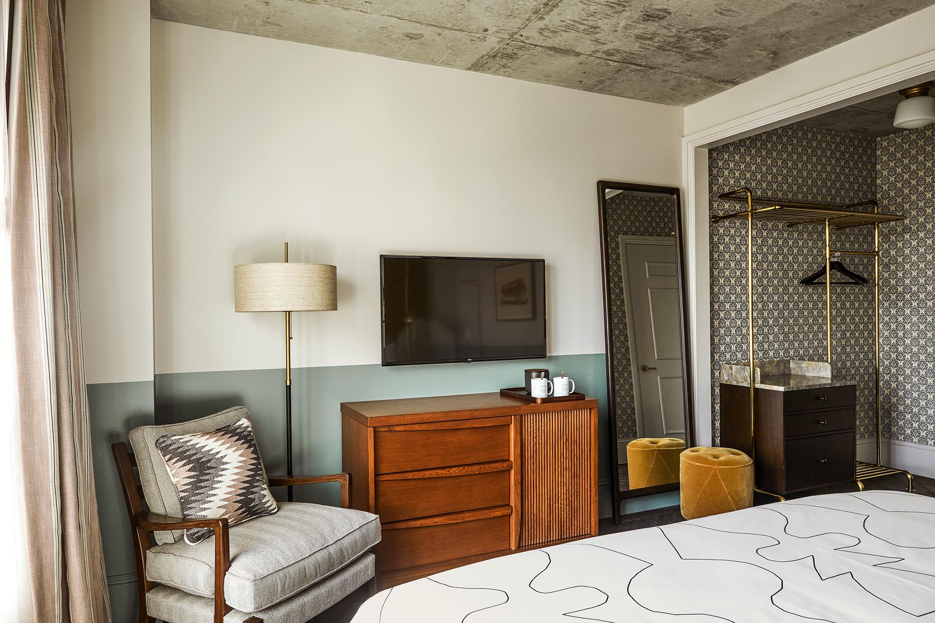 The room includes a lounge chair, drawers, floor lamp, TV on the wall, full length mirror and a hanging area for clothes