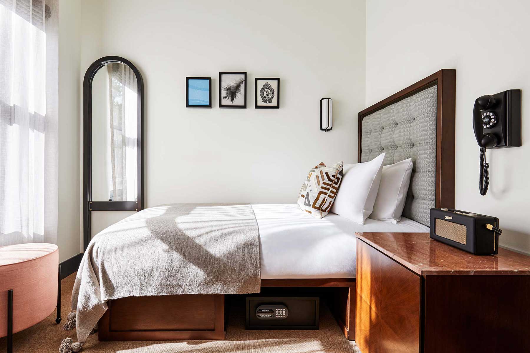 A single bed sits against a wall, in a compact room which has a full length mirror, footstool and a safe under the bed