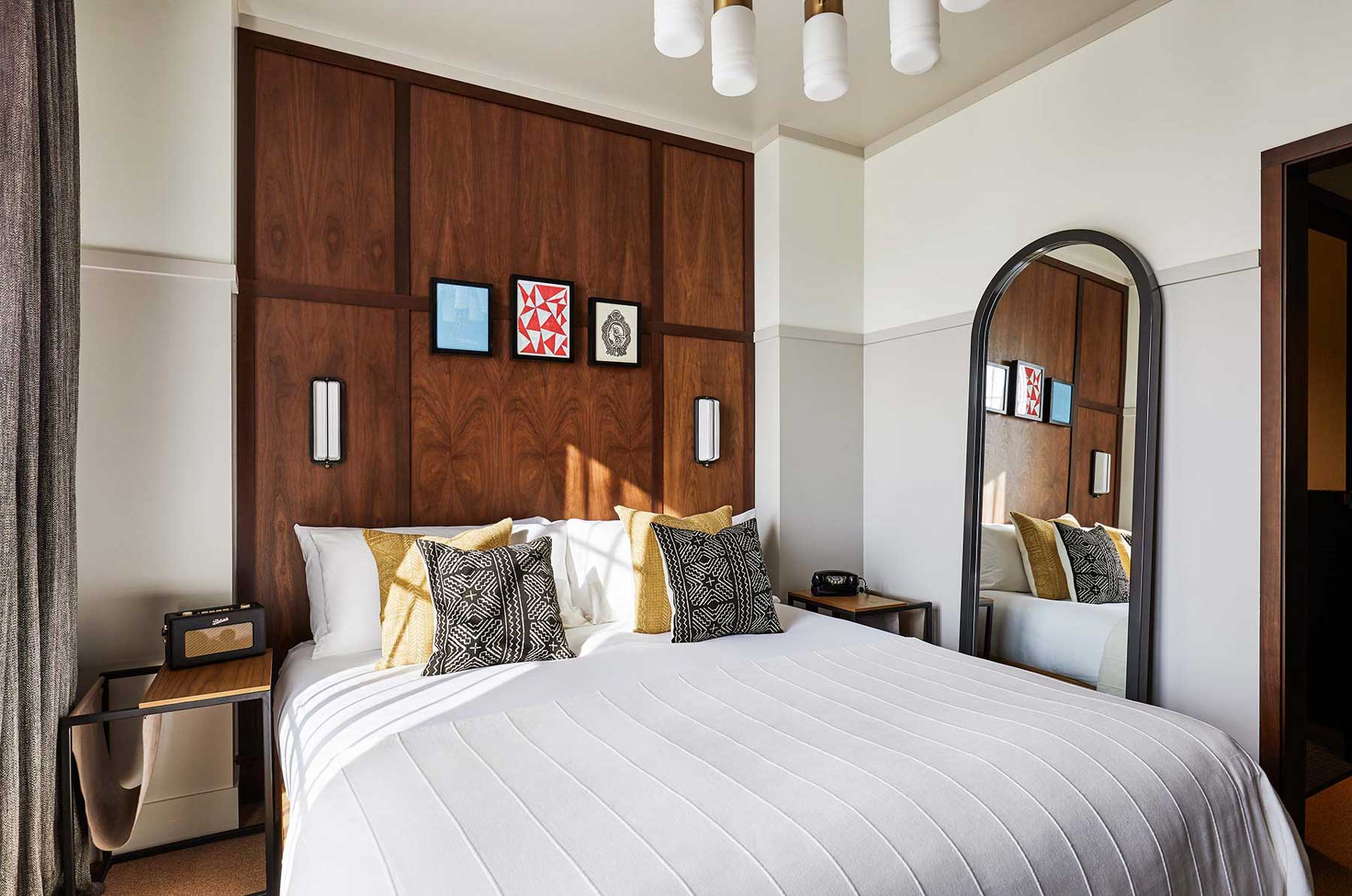 A full length mirror leans against the wall beside the bed, which has white linen and four patterned cushions on top