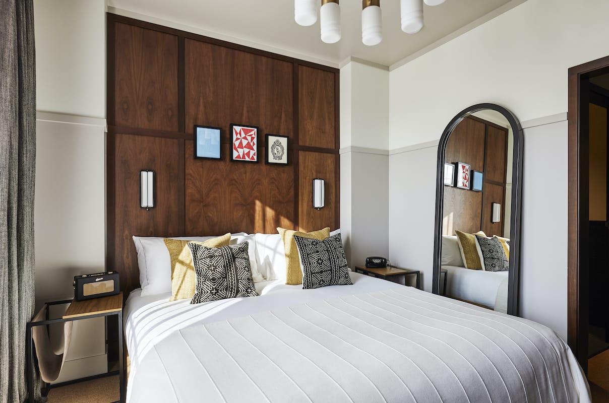 A Queen sized bed with crisp white sheets and cushions, with a full length mirror leaning on the wall beside it