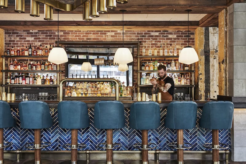 A man works in a bar with an exposed brick back, bright blue tiling and blue bar stools