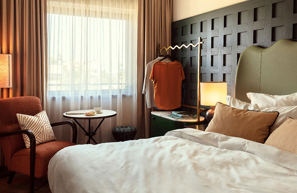 The hotel room includes an area to hang clothes, a lounge chair and table, and lamps