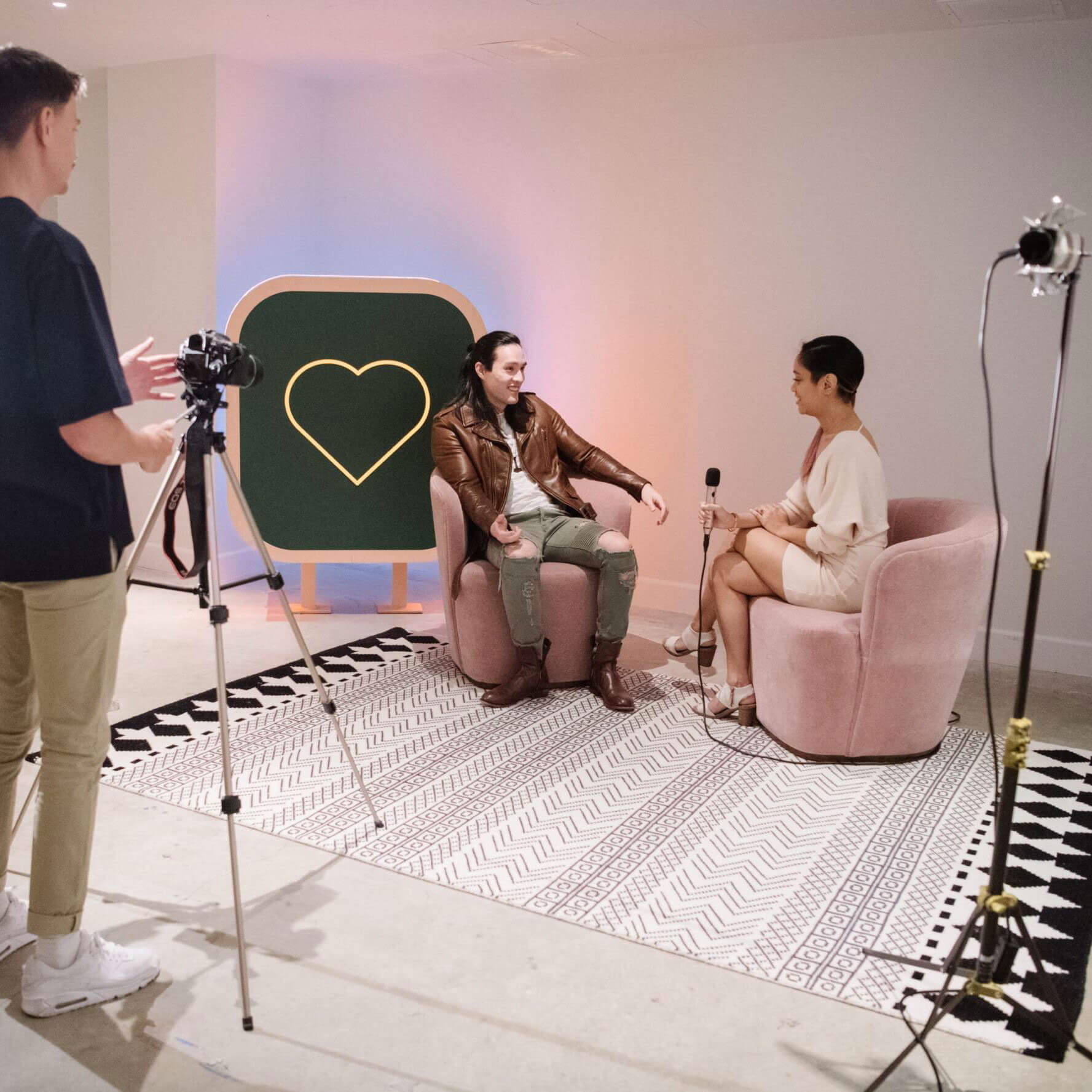 Man with a camera on a tripod films two people having an interview on pink seats