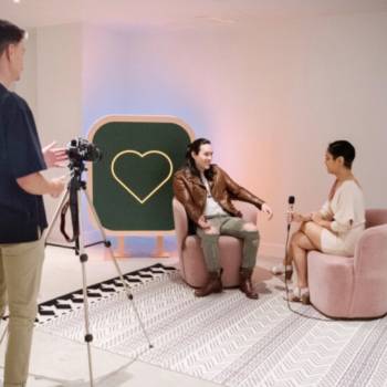 Man with a camera on a tripod films two people having an interview on pink seats