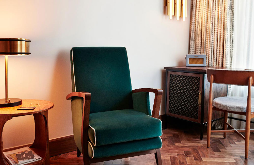 A dark green velvet lounge chair beside a small table. The floor is wooden. There is a desk and chair in the corner.