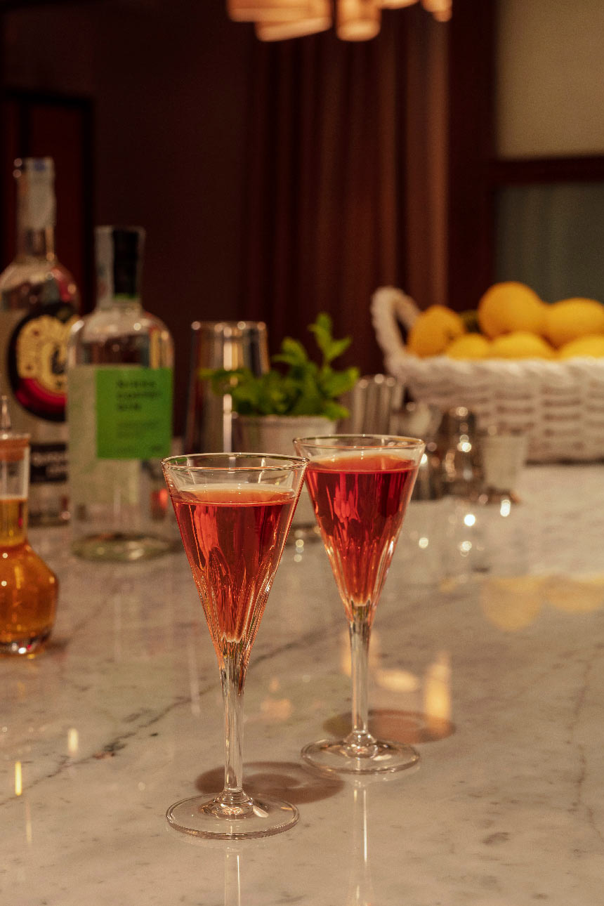 Two glasses of a rose coloured liquid in front of bottles of drink and a basket full of lemons