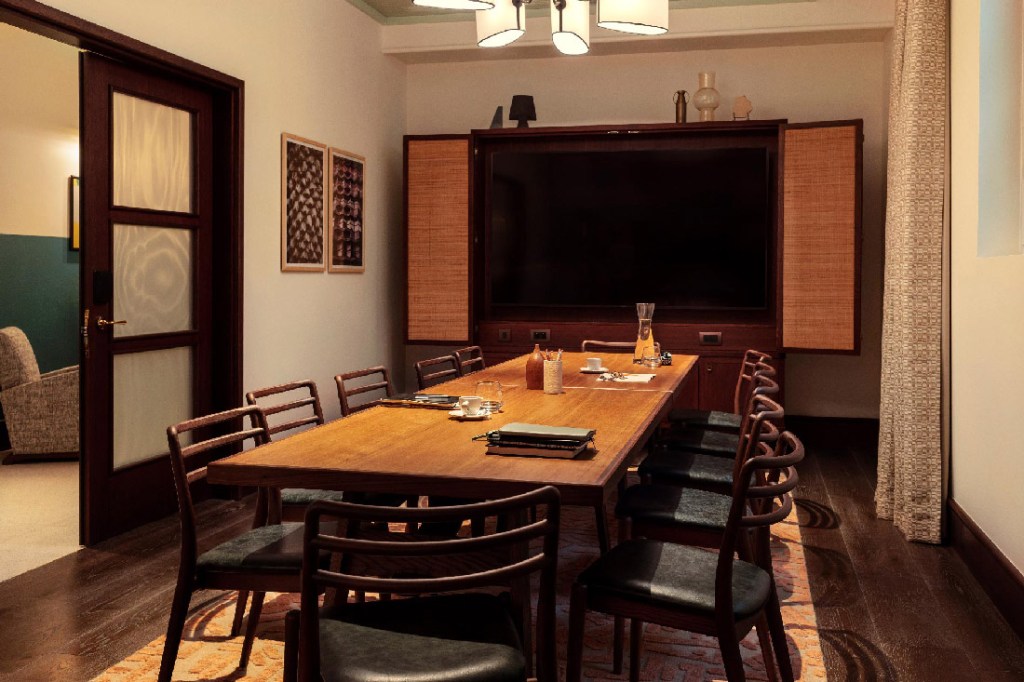 Meeting room with long table and 12 chairs, with large TV screen at the end of the table. Water, books, pencils and a laptop are on the table.