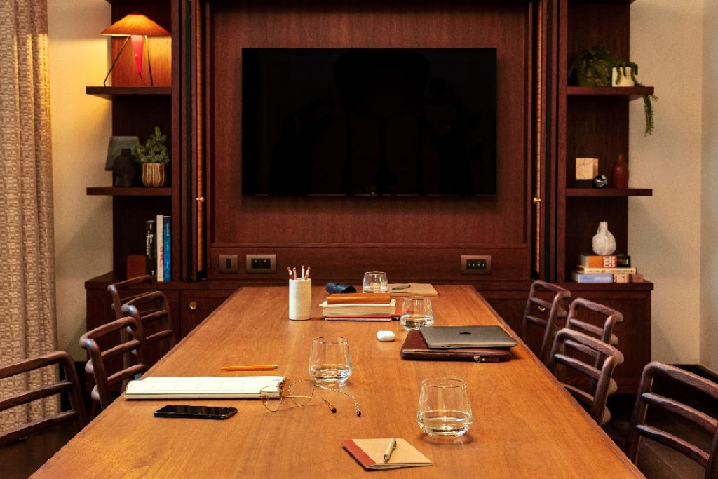 Meeting room with long table and 8 visible chairs, with large TV screen at the end of the table. Water, books, pencils and a laptop are on the table.