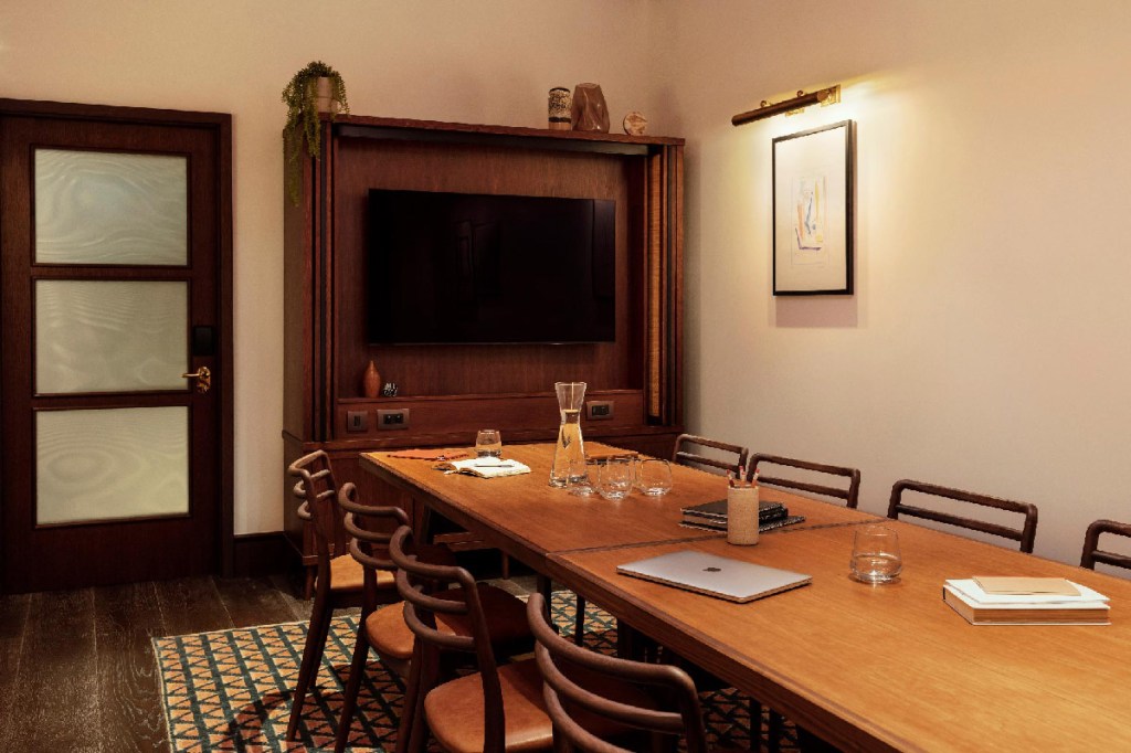 Meeting room with long table and 8 chairs, with large TV screen at the end of the table. Water, books, pencils and a laptop are on the table.