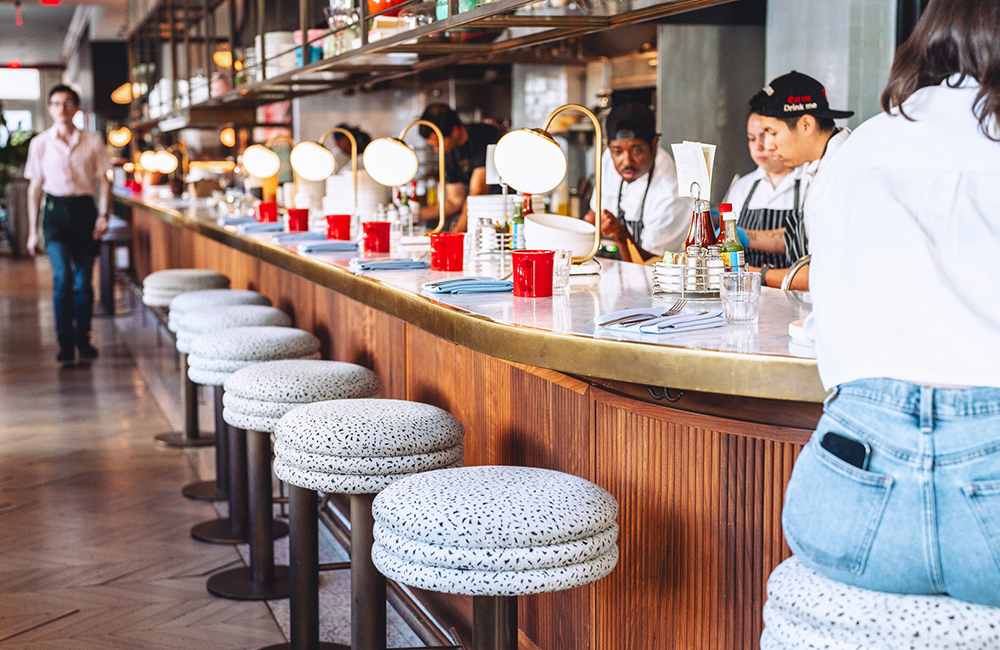 Stools line the bar with chefs preparing food and drinks behind
