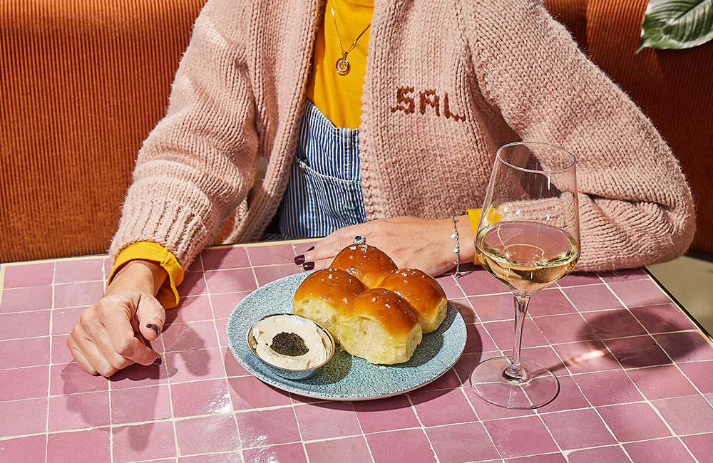 A diner in a pink cardigan sits with a glass of white wine and a plate of K'Far food.