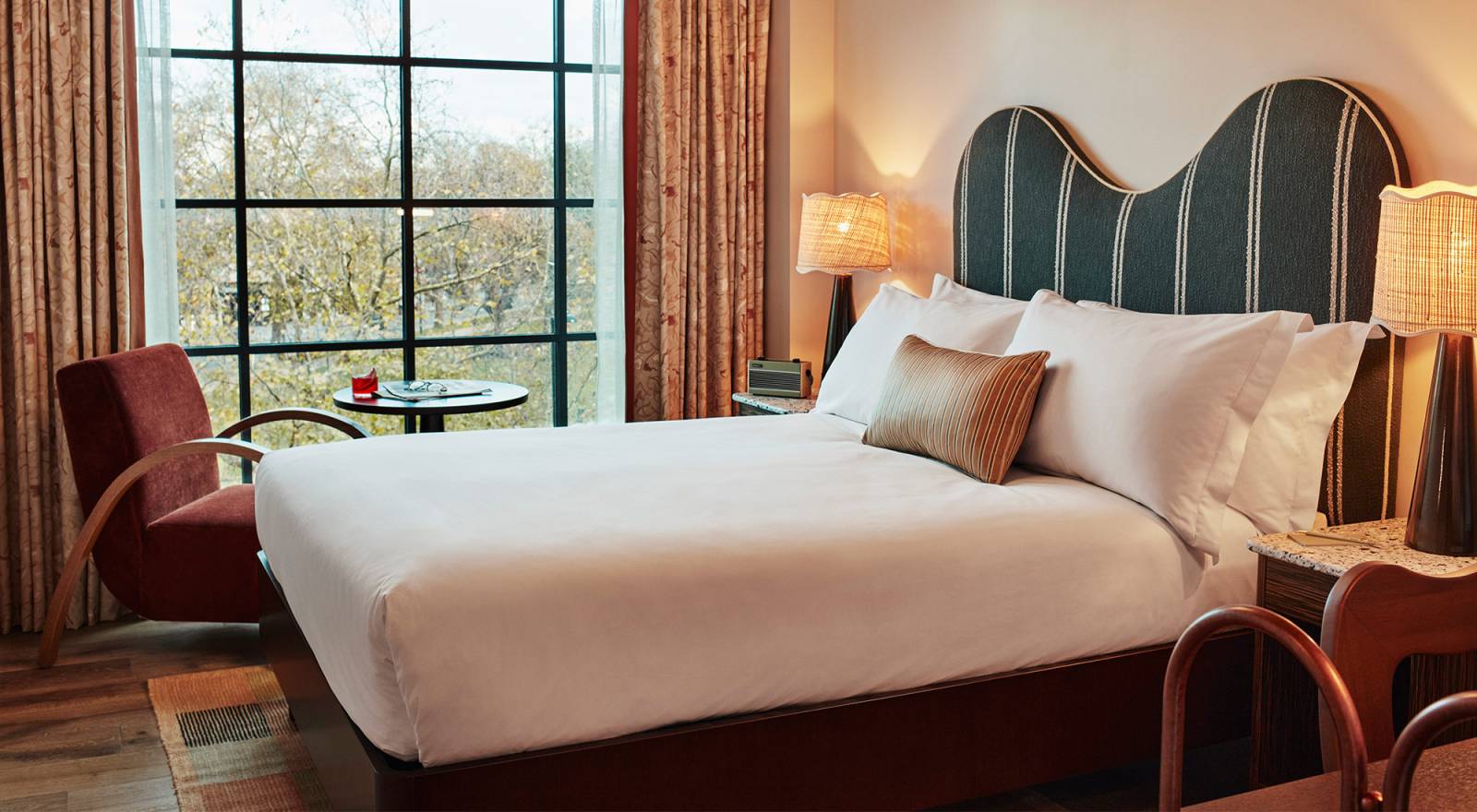 Interior of a room at our West London Hoxton hotel with plush double bed, comfortable pillows, bedside lamps, table, armchair and floor to ceiling window with bright view.
