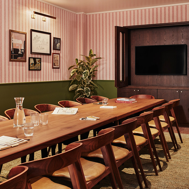 A meeting space available to hire at The Hoxton in Shepherd's Bush with places arranged on a long wooden table with a natural plant and a video screen visible.
