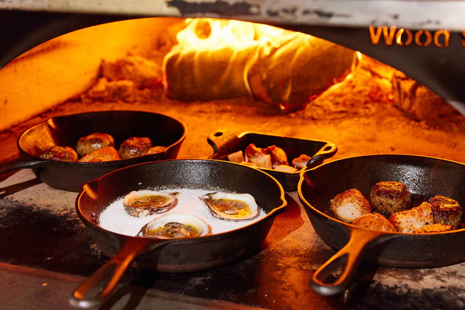 Pans of scallops rest in an open oven in which a wood fire burns.