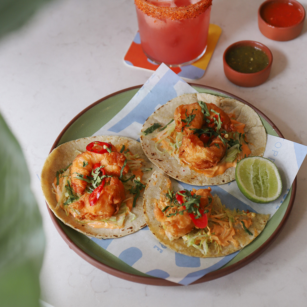 Tasty prawn tacos with a red cocktail seen behind.