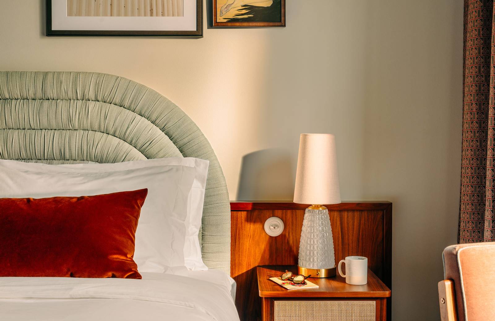 Sunlight crosses a lamp on a bedside table next to a comfy bed with red pillows
