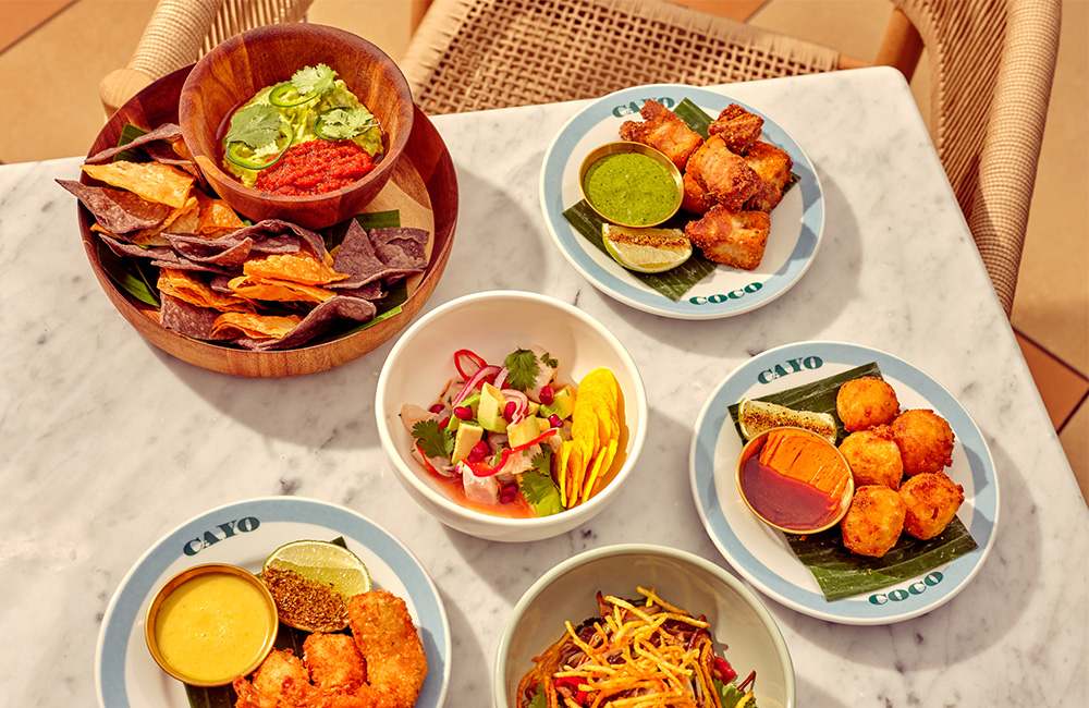 Overview of mixed dishes of food, from snacks to main dish, served on a marble table and raffia chair next.