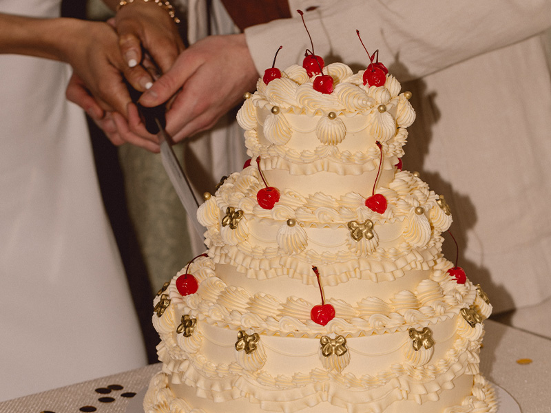 couple holding cake knife and cutting into a 3-tier white decorated wedding cake with cherries.