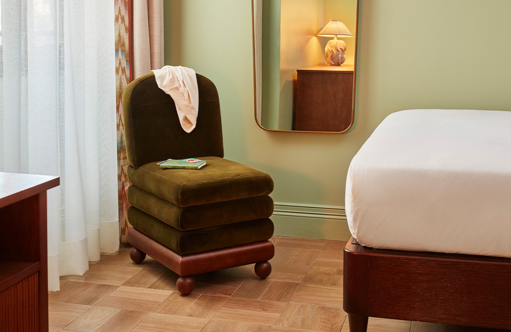 End of a bed with a dark wood frame, a single green velvet chair beside an open window, and a mirror reflecting a desk.