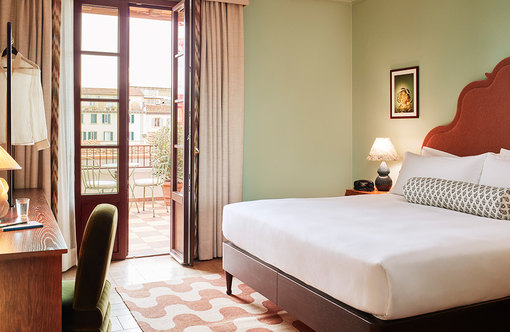 Large window allowing natural sunlight to illuminate a pink upholstered bed frame, with a green chair tucked under a desk and large wooden doors leading to a terrace with an outdoor seating area.