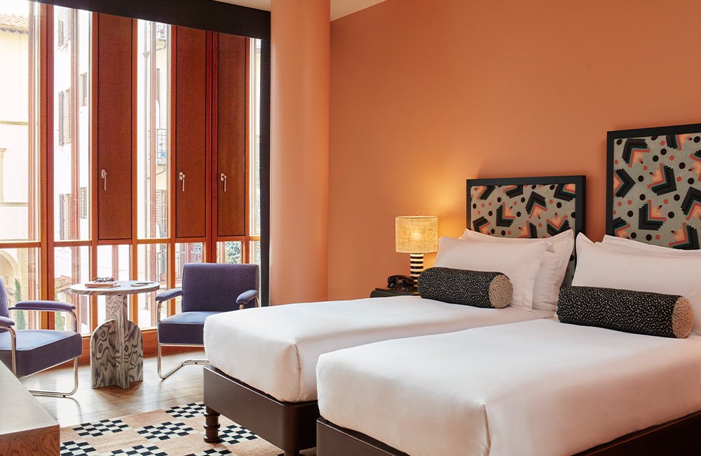 Two single beds with large printed bolsters on each, accompanied by black and white side lamps on separate bedside tables. A large window with a seat beside it fills the room with natural light.