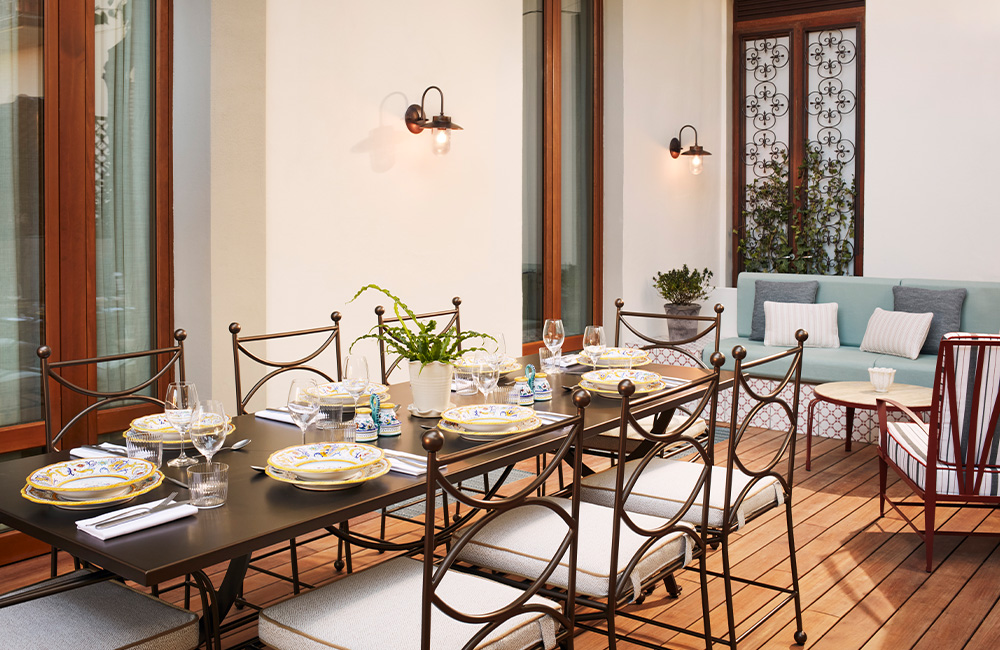 Spacious house terrace with a wooden deck, featuring a long table set for 8 people. Behind it, next to the interior wall, is a large teal outdoor sofa with a coffee table.