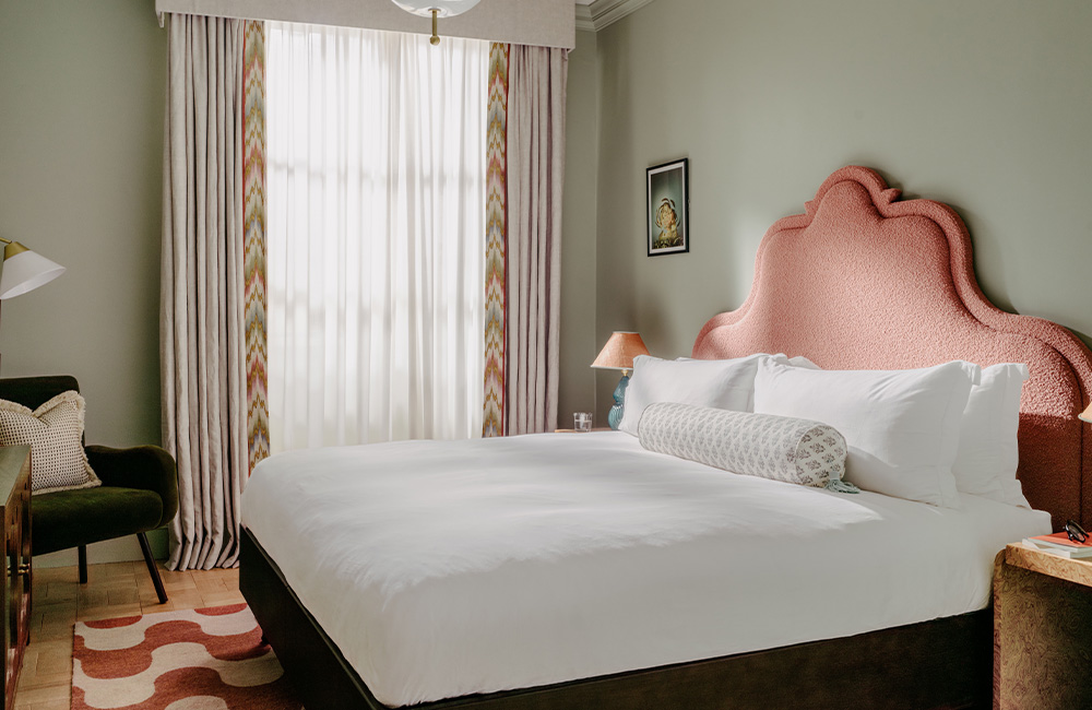 Overview of a spacious hotel room featuring a large king-size bed, an armchair, a bedside table, and a large window allowing natural light to fill the space