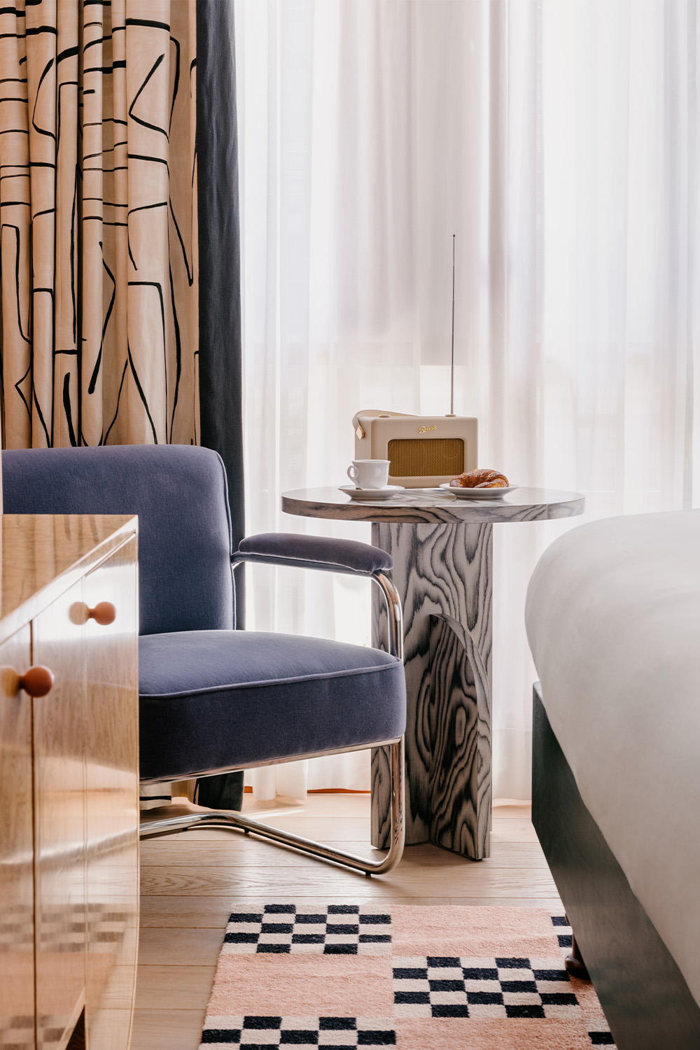 End of the bed featuring a blue velvet armchair next to a marble side table, with natural light streaming in from a large window