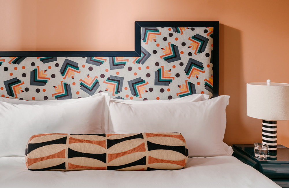Headboard with a bright, dynamic pattern, large fluffy pillows, and a black and white lamp beside it.