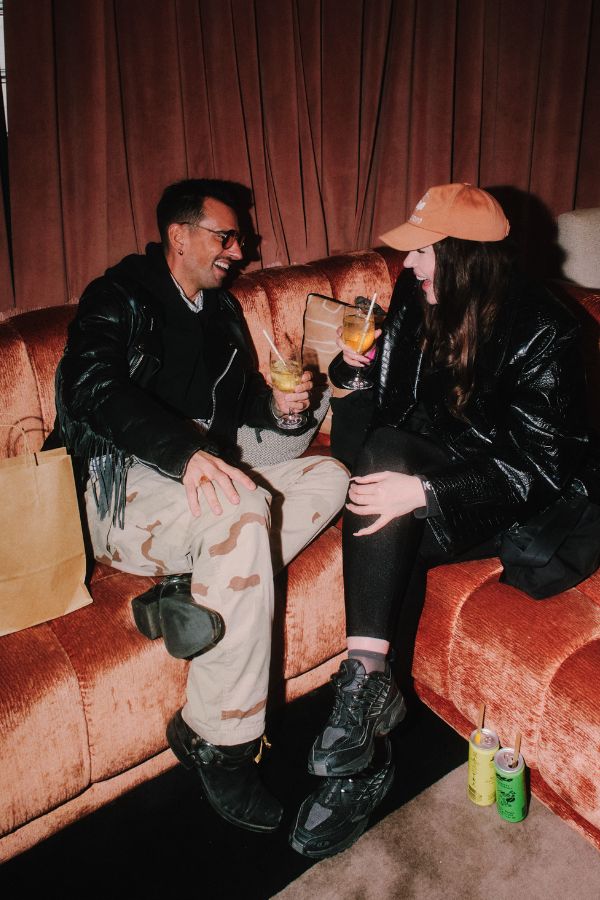 Two people sitting on a pink velvet sofa talking and laughing while drinking their colourful beverages.